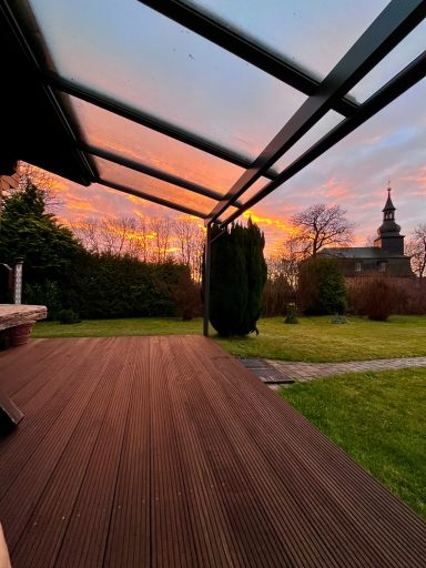 Sonnenuntergang mit bunten Wolken über einem Garten und einem Kirchturm im Hintergrund.