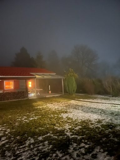 Ein Holzhaus mit rotem Dach, umgeben von Schnee und Nebel, warm beleuchtet.