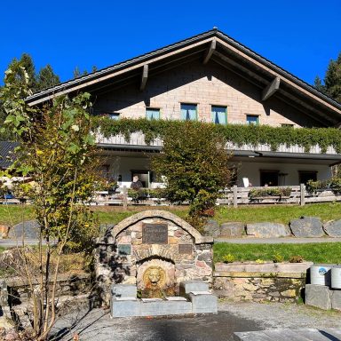 Werraquell Hütte Ein Haus mit Balkon, umgeben von Bäumen und einem Brunnen im Vordergrund.