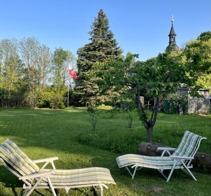 Zwei Liegen im Garten unter Bäumen, im Hintergrund eine Kirche und ein Flaggenmast.